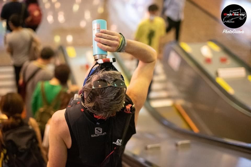 Back of Rich Cowler's head wearing a Salomon cap, water bottle visible over his shoulder on a sunny trail