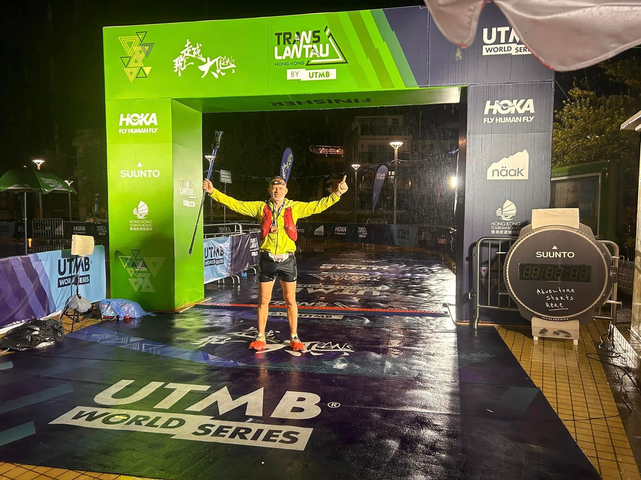 Rich Cowler crossing the UTMB Trans Lantau finish line at night, trekking poles raised in triumph