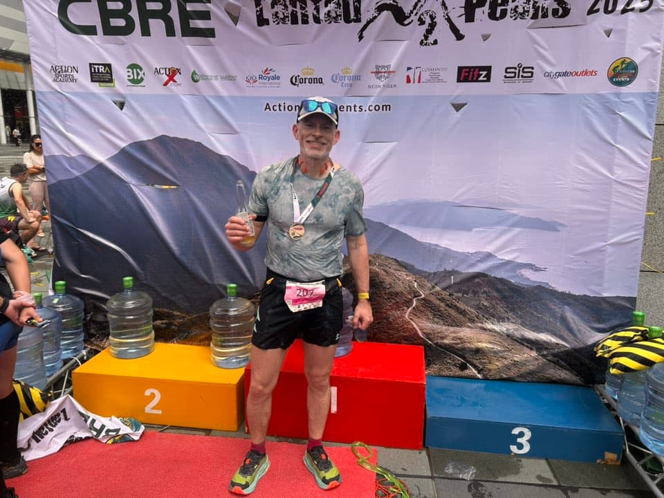 Rich Cowler on the podium at CBRE Lantau 2 Peaks 2023, holding a medal and champagne